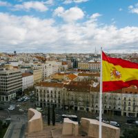 Drone view of Spanish city with aged buildings and national flag under cloudy blue sky